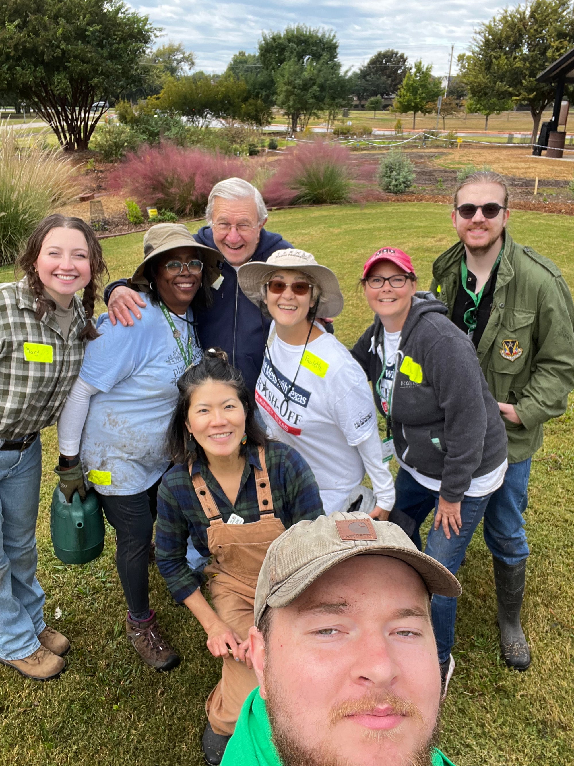A group of seven volunteers posing together outdoors in a garden setting, wearing matching yellow name tags and casual work clothes. The photo is a selfie-style group shot taken on a grassy lawn with plants and trees in the background.